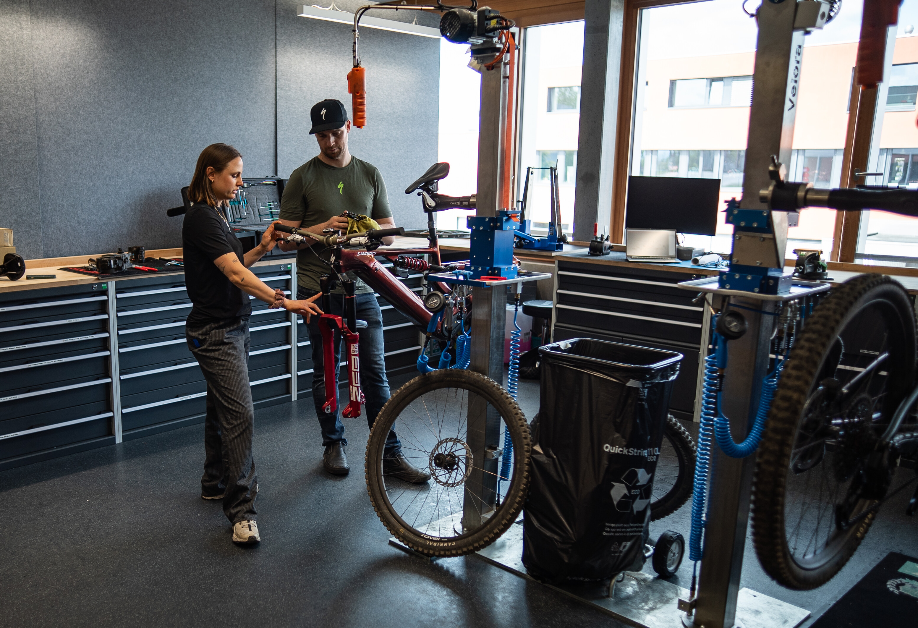 Sofia Wiedenroth steht mit einem Fahrrad-Techniker in einer Werkstatt an einem montierten Fahrrad. Sofia zeigt auf ein Bauteil, während der Fahrrad-Techniker danebensteht und etwas in den Händen hält.