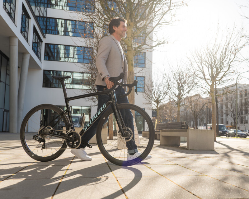 Ein Mann schiebt ein Rennrad der Marke Specialized auf einem sonnigen Platz vor einem modernen Bürogebäude.