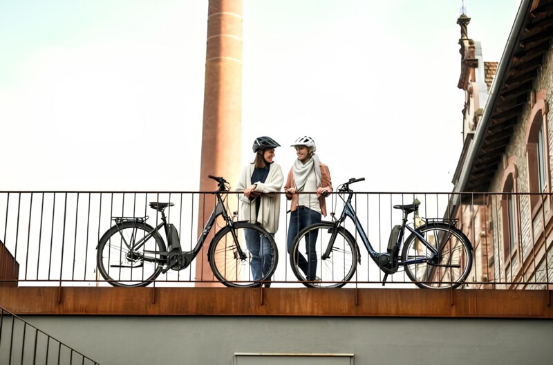 Zwei Frauen mit Fahrradhelmen lehnen an einem Geländer, neben ihren Fahrrädern, mit einem Backstein-Gebäude und Schornstein im Hintergrund.