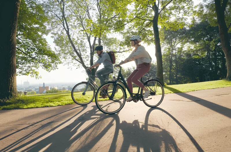 Zwei Personen fahren bei sonnigem Wetter mit Fahrrädern auf einem Weg durch einen Park. Die langen Schatten der Bäume und der Radfahrer erstrecken sich über den Boden, und im Hintergrund sind Bäume und eine Aussicht auf die Stadt sichtbar.