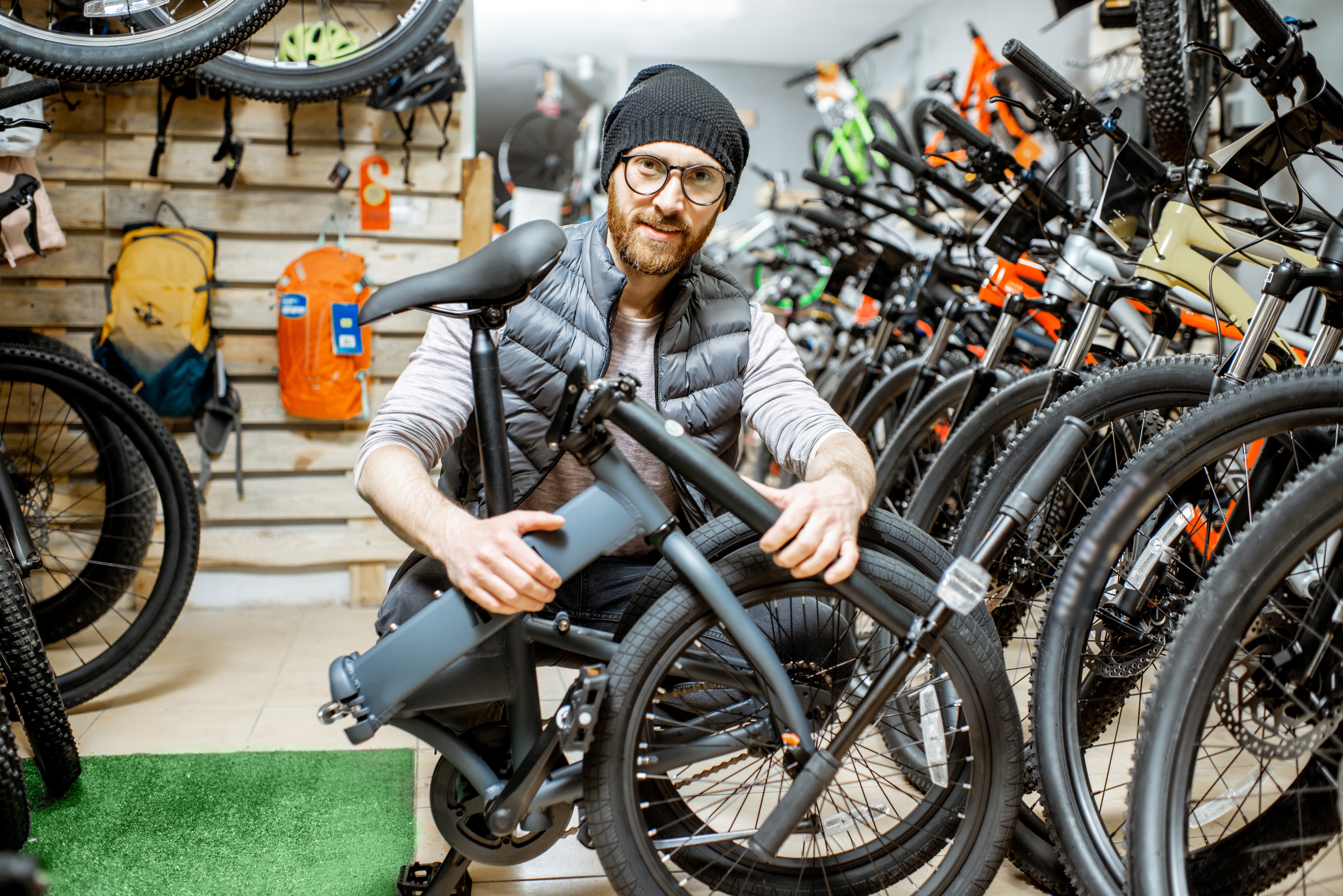 Ein Mann in Mütze und Brille kniet in einem Fahrradladen und hält ein zusammengefaltetes Klapprad.