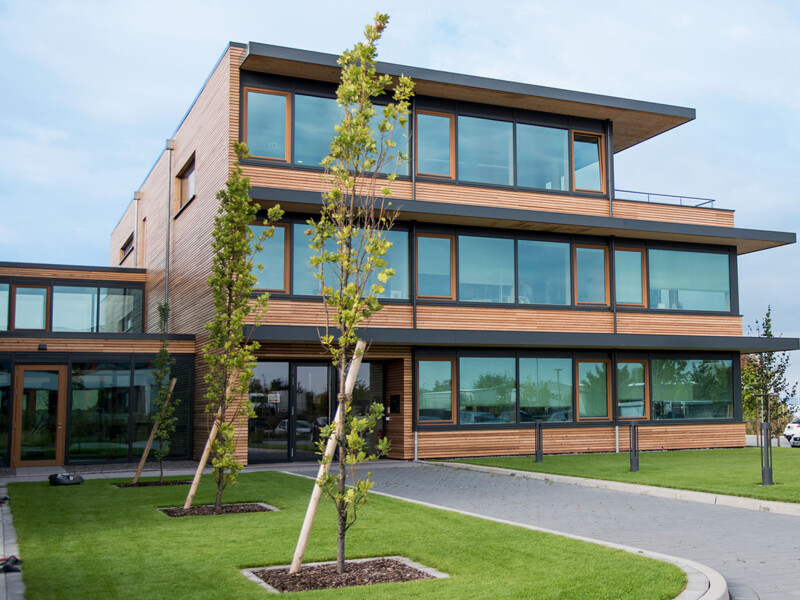 Modernes Gebäude mit Holzverkleidung, großen Glasfenstern und grünem Rasen im Vordergrund.