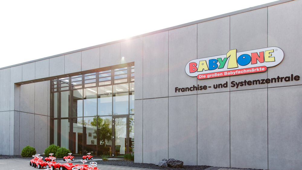 Moderne Gebäudefassade mit dem farbenfrohen Logo von "BabyOne" und dem Schriftzug "Franchise- und Systemzentrale". Vor dem Eingang stehen mehrere rote Spielzeugautos.