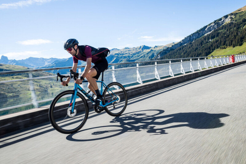 Ein Radfahrer in sportlicher Kleidung fährt über eine Brücke mit Bergen im Hintergrund bei sonnigem Wetter.