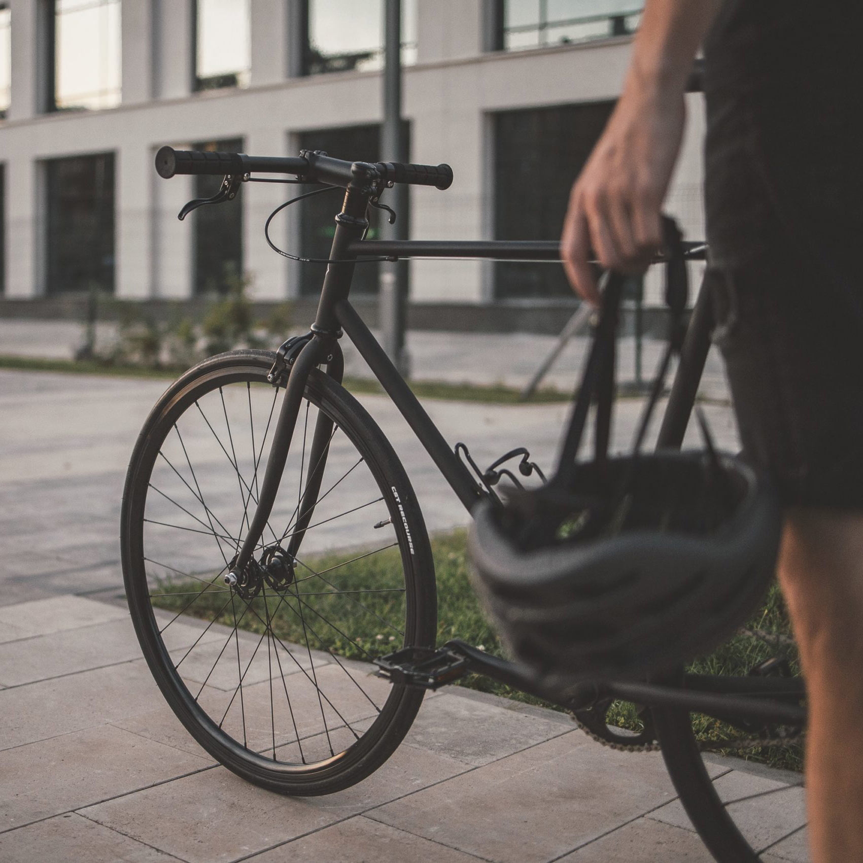 Eine Person hält einen Fahrradhelm in der Hand, während sie neben einem schwarzen Fahrrad steht. Im Hintergrund ist ein modernes Gebäude mit großen Fenstern zu sehen.