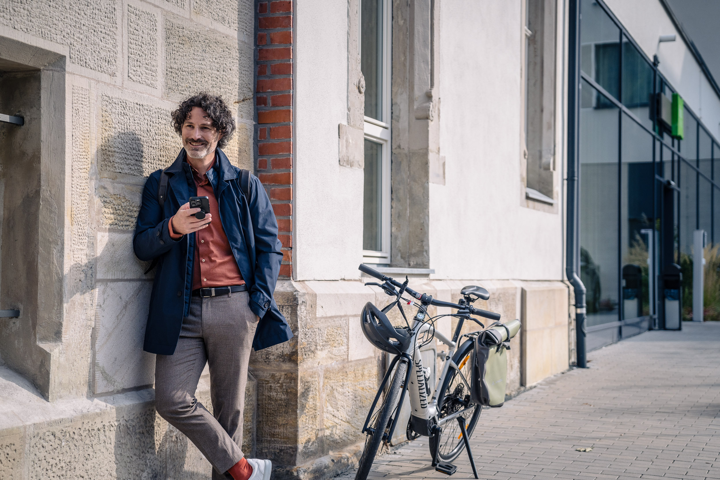 Ein Mann lehnt lächelnd an einer Gebäudewand und schaut auf sein Smartphone, während neben ihm ein Fahrrad am Gehweg steht.