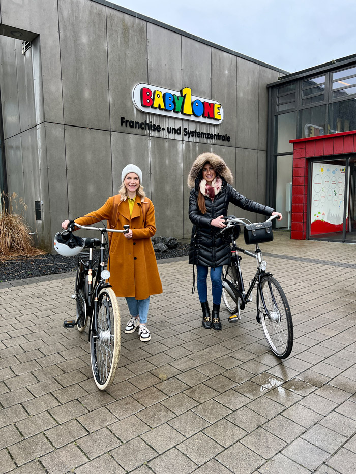 Zwei Frauen stehen mit Fahrrädern vor dem Gebäude der BabyOne Franchise- und Systemzentrale.