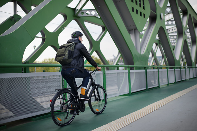 Ein Mann mit Helm und Rucksack fährt mit einem Fahrrad über eine Brücke mit grünem Stahlgerüst. Der Boden der Brücke ist grün gestrichen, und die Struktur besteht aus diagonalen und geometrischen Elementen.