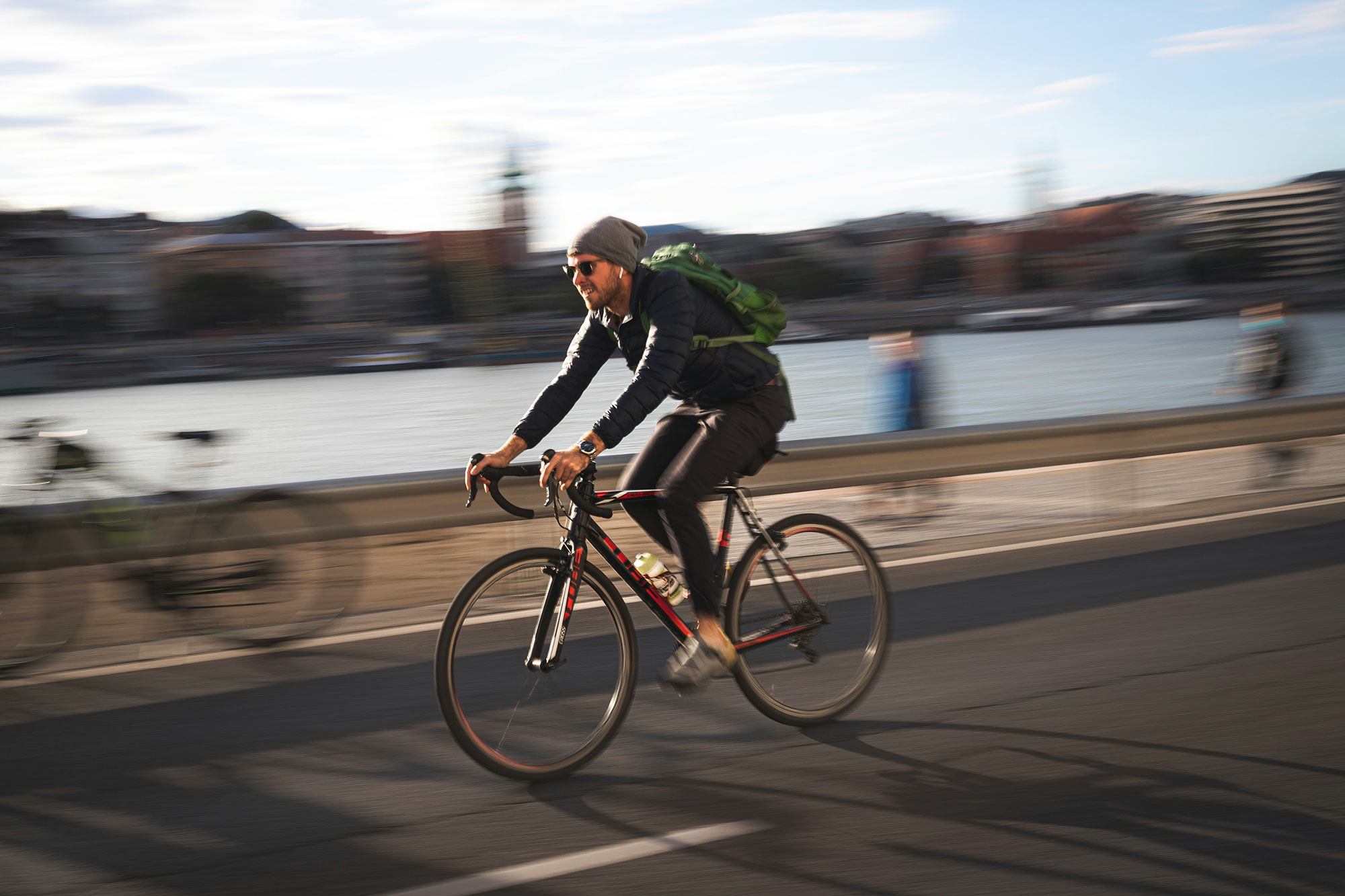 Ein Radfahrer mit Rucksack fährt zügig am Wasser entlang, der Hintergrund ist durch Bewegungsunschärfe verwischt.