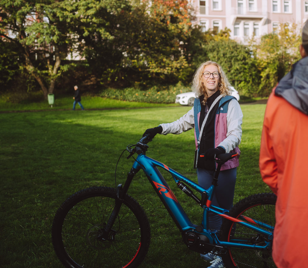 Eine Frau mit lockigem Haar und Brille steht auf einer grünen Wiese neben einem blauen E-Bike. Im Hintergrund sind Bäume und Gebäude zu sehen. Sie trägt eine bunte Jacke und lächelt zu einer Person rechts im Bild.