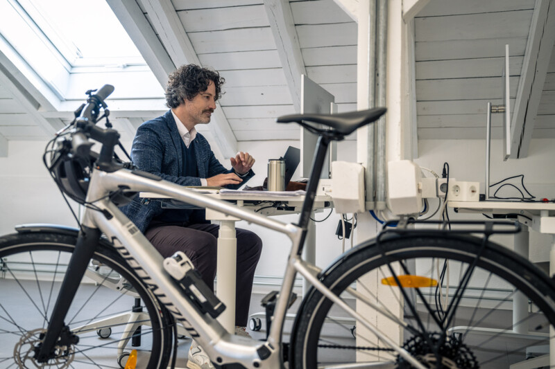 Ein Mann arbeitet am Laptop an einem Schreibtisch im hellen Dachgeschossbüro, während im Vordergrund ein Fahrrad steht.