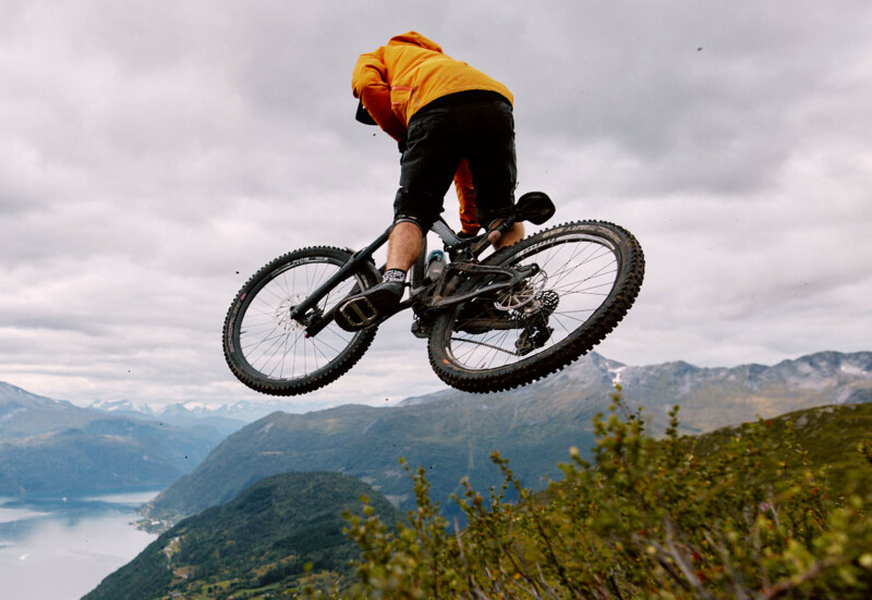 Ein Mountainbiker in einem orangefarbenen Oberteil springt mit seinem Fahrrad in die Luft, während im Hintergrund eine malerische Berglandschaft mit einem See zu sehen ist.