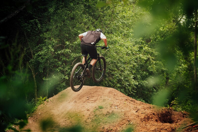 Ein Mountainbiker springt auf einem Waldtrail über einen Erdhügel, eingerahmt von dichtem Grün.