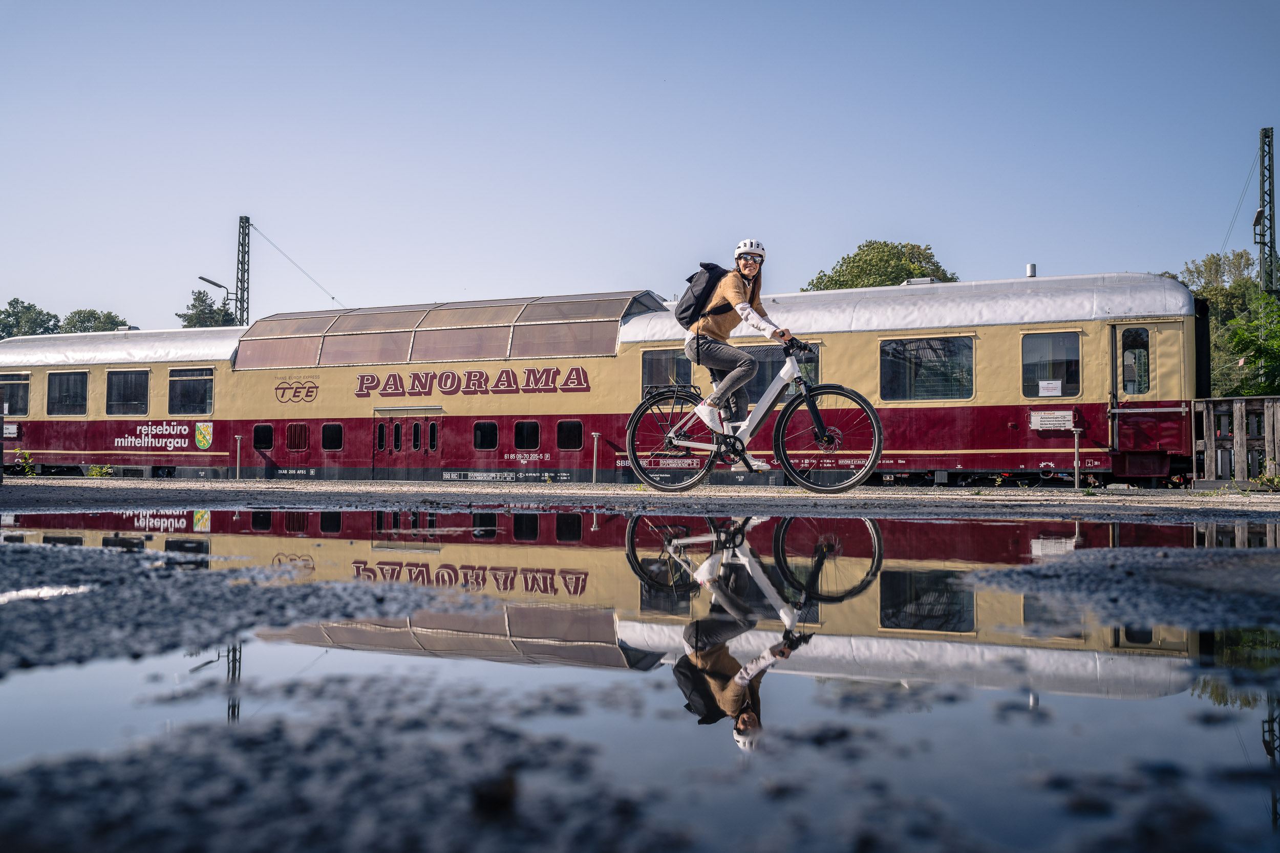 Eine Radfahrerin mit Helm fährt an einem historischen „Panorama“-Zug vorbei, der sich in einer Pfütze im Vordergrund spiegelt.