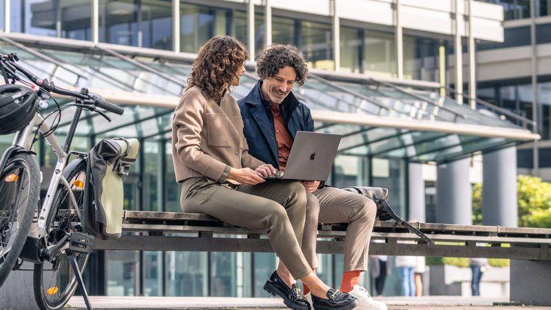 Zwei Kolleg:innen sitzen auf einer Bank vor einem modernen Bürogebäude und schauen auf einen Laptop neben einem geparkten Fahrrad.