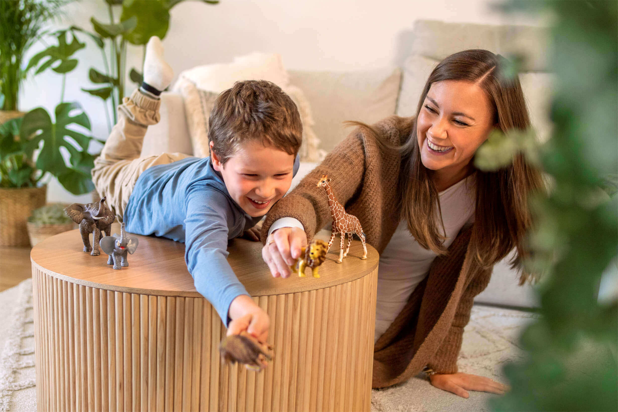 Eine lachende Frau spielt mit einem kleinen Jungen, der auf einem runden Holztisch liegt. Beide bewegen kleine Spielzeugtiere wie Elefanten, Löwen und Giraffen. Im Hintergrund sind grüne Zimmerpflanzen und ein Sofa zu sehen.