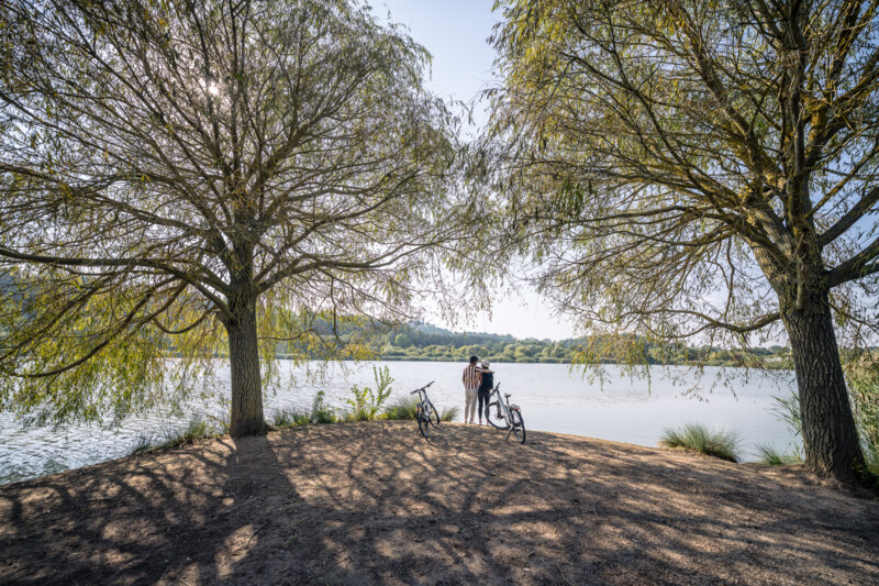 Zwei Personen stehen am Ufer eines Sees zwischen zwei großen Bäumen, neben ihnen lehnen Fahrräder im Schatten.