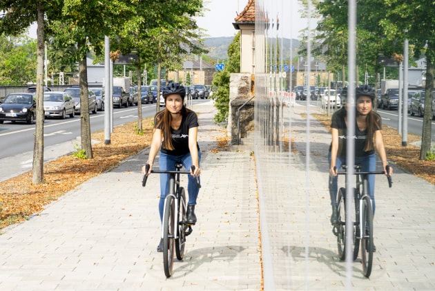 Eine Frau fährt auf einem Fahrrad auf einem Gehweg entlang, und ihre Spiegelung ist auf der Glaswand rechts neben ihr zu sehen.