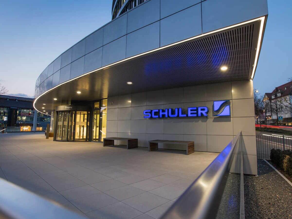 Beleuchteter Eingang eines modernen Gebäudes bei Dämmerung, mit dem Schriftzug 'Schuler' in blau an der Fassade.