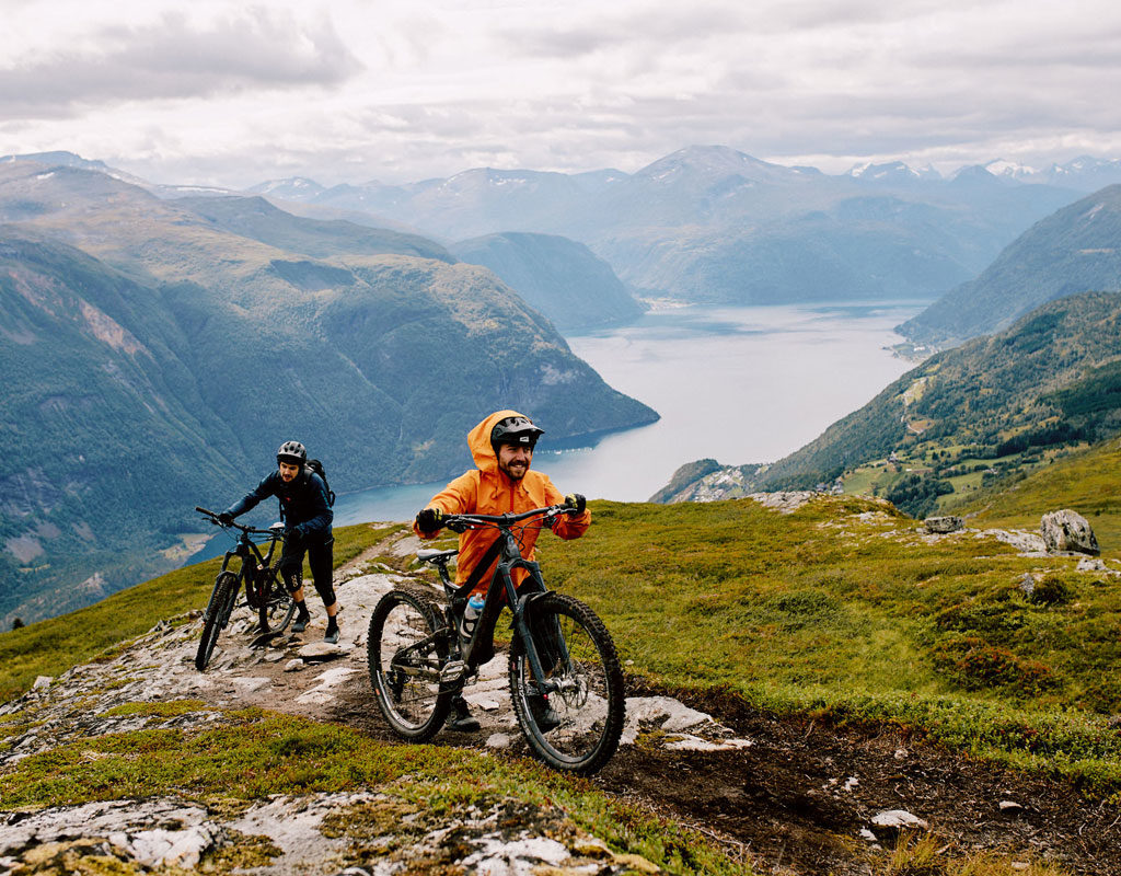Zwei Mountainbiker schieben ihre Fahrräder einen steilen Bergpfad hinauf, mit einem weiten Blick auf einen See und umgebende Berge im Hintergrund.