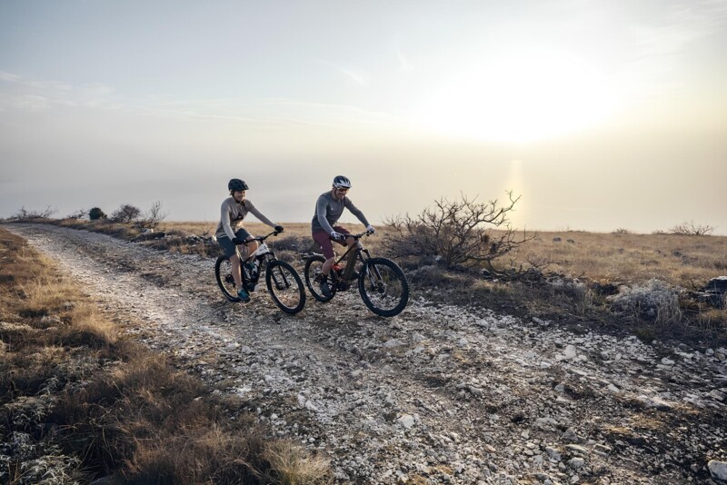Zwei Personen fahren mit Mountainbikes auf einem Schotterweg bei Sonnenaufgang oder Sonnenuntergang in einer offenen, naturbelassenen Landschaft.