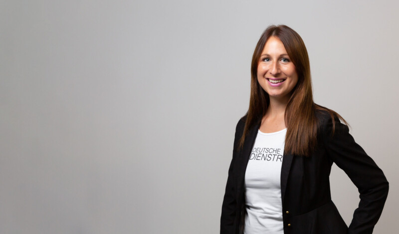 Eine Frau in einem schwarzen Blazer mit einem weißen T-Shirt, auf dem "DEUTSCHE DIENSTRAD" steht, vor neutralem grauem Hintergrund.