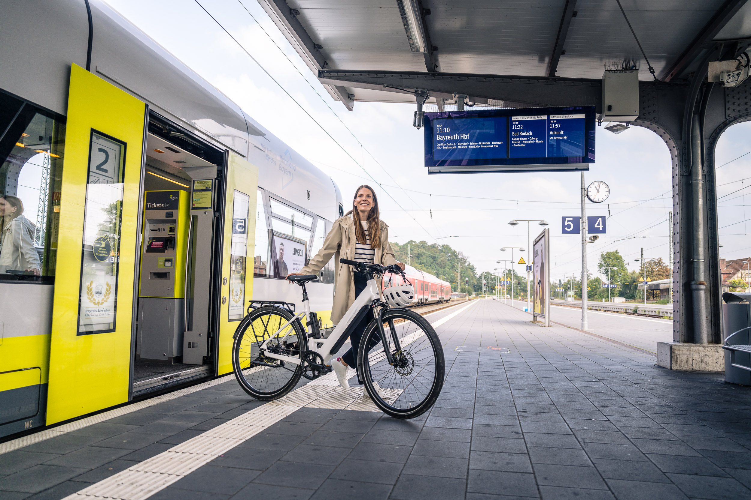 Eine Frau steht mit einem Fahrrad am Bahnsteig neben einem gelben Zug mit offener Tür, darüber hängen Anzeigetafeln und Gleisnummern.